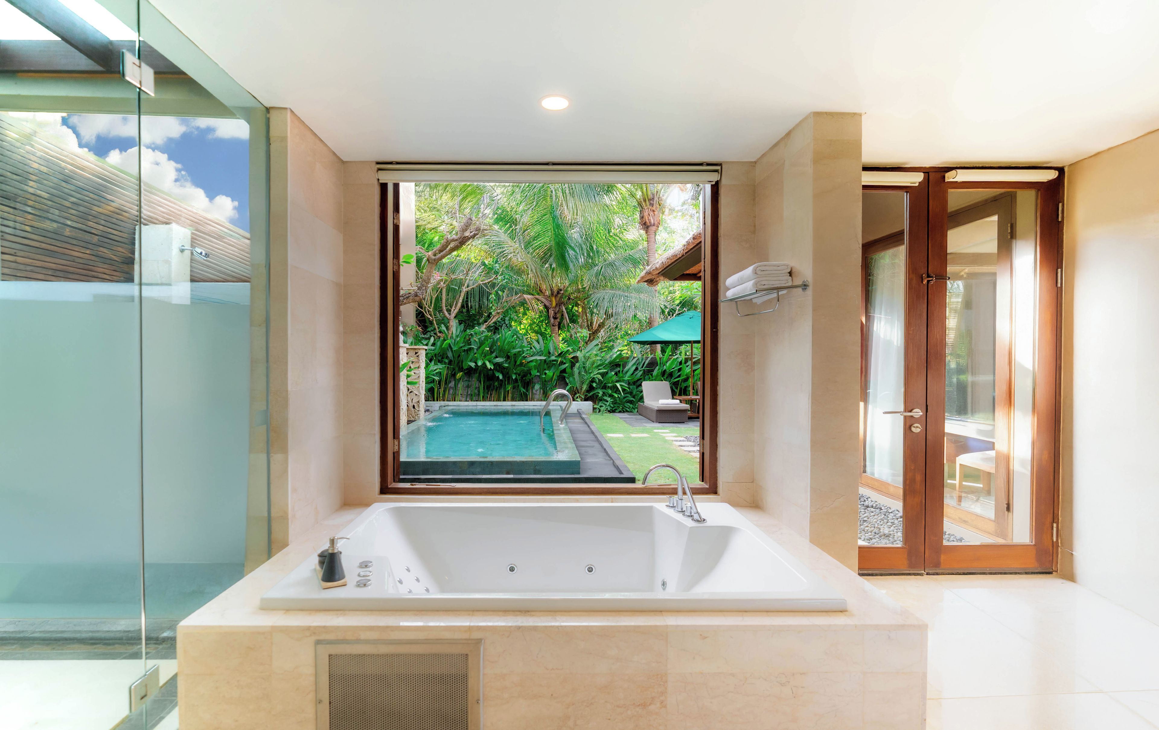 A modern bathroom with a large bathtub, a glass shower enclosure, and a view of a pool and greenery outside.