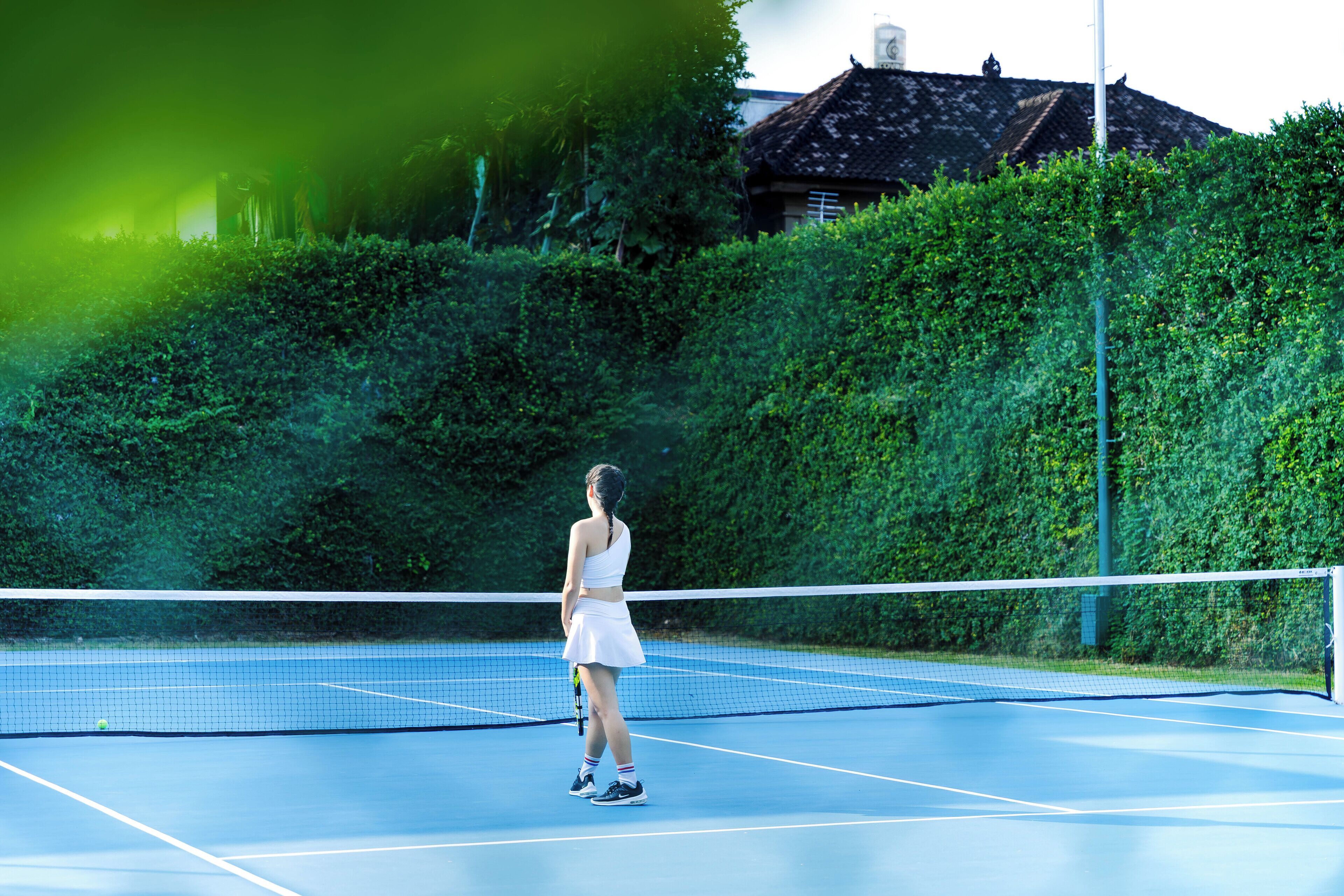A tennis court with a net, surrounded by greenery and a building in the background.