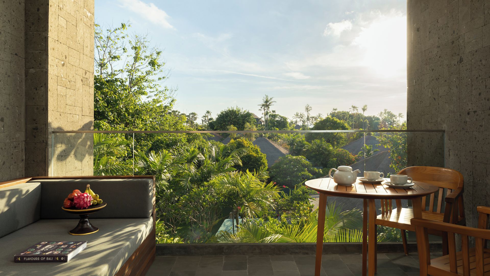 A balcony with a view of a lush garden, a round wooden table with a teapot and cups, and a sofa with a fruit bowl.