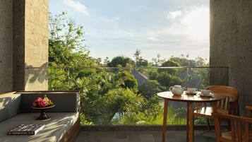 A balcony with a view of a lush garden, a round wooden table with a teapot and cups, and a sofa with a fruit bowl.