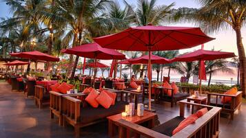 An outdoor seating area with wooden furniture, red umbrellas, and palm trees.
