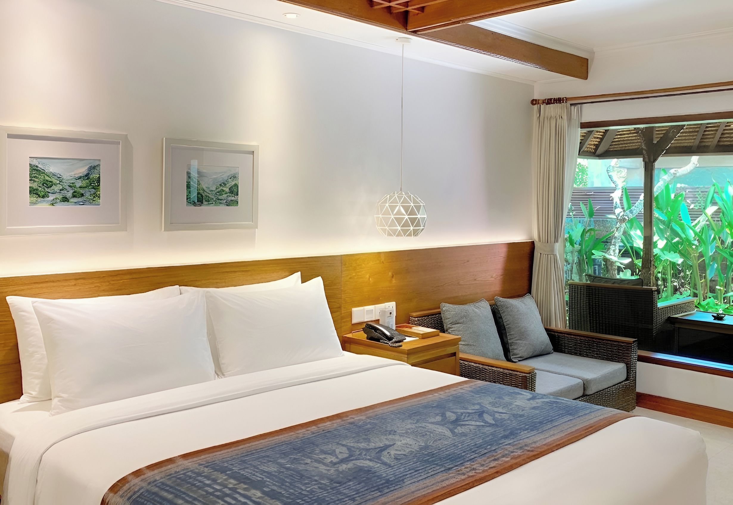A hotel room with a large bed, a bedside table, a chair, and a window with a view of greenery.