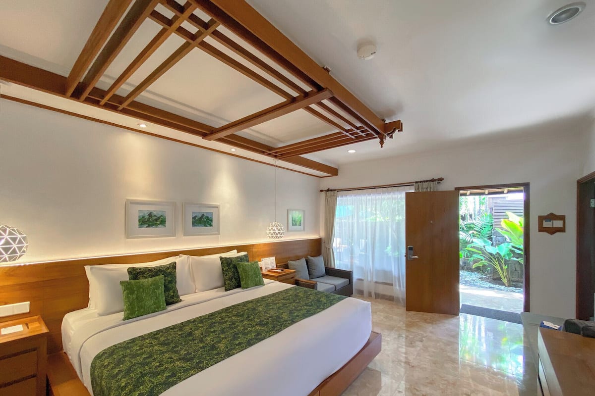 A hotel room with a large bed, wooden headboard, and a view of greenery through the door.