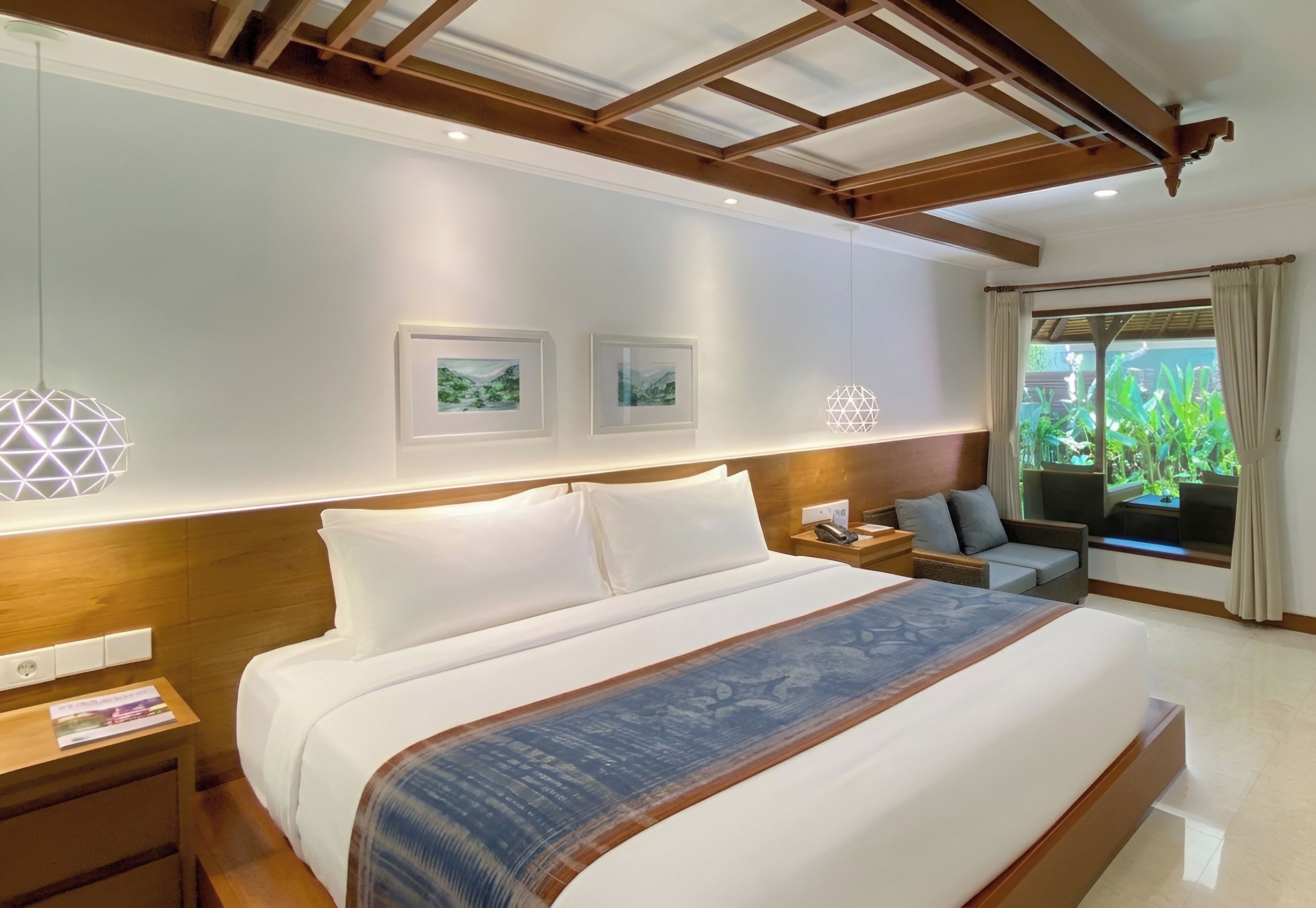 A hotel room with a large bed, two bedside tables, a nightstand, a sofa, a window with curtains, and two framed pictures on the wall.