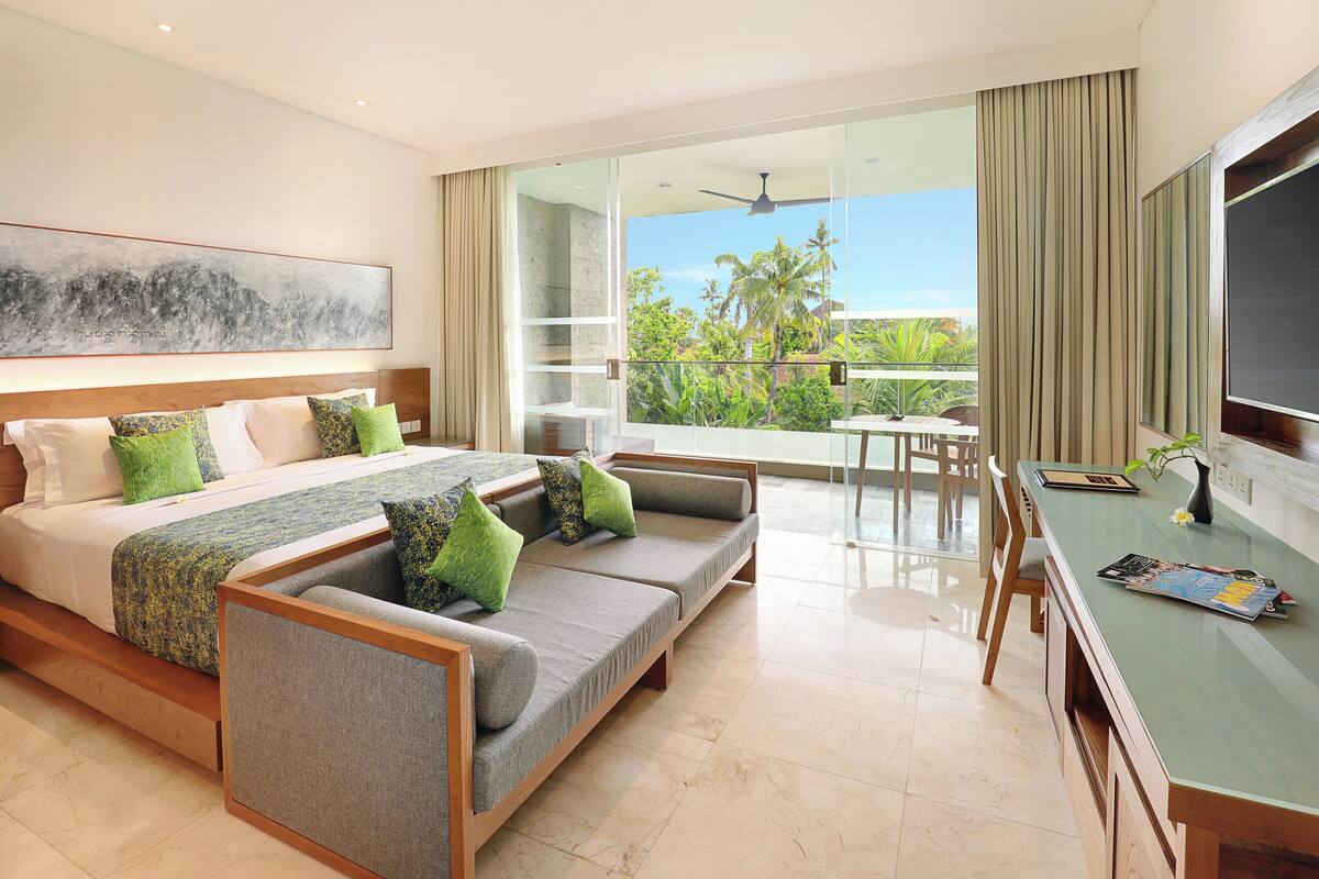 A modern hotel room with a large bed, a sofa, a dining area, and a balcony with a view of palm trees.