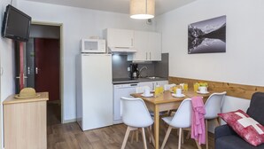 Cocina compacta con armarios blancos, microondas y cafetera. Hay una mesa de comedor con sillas y un banco de madera. Un televisor está montado en la pared, y se observa un cuadro de una montaña.