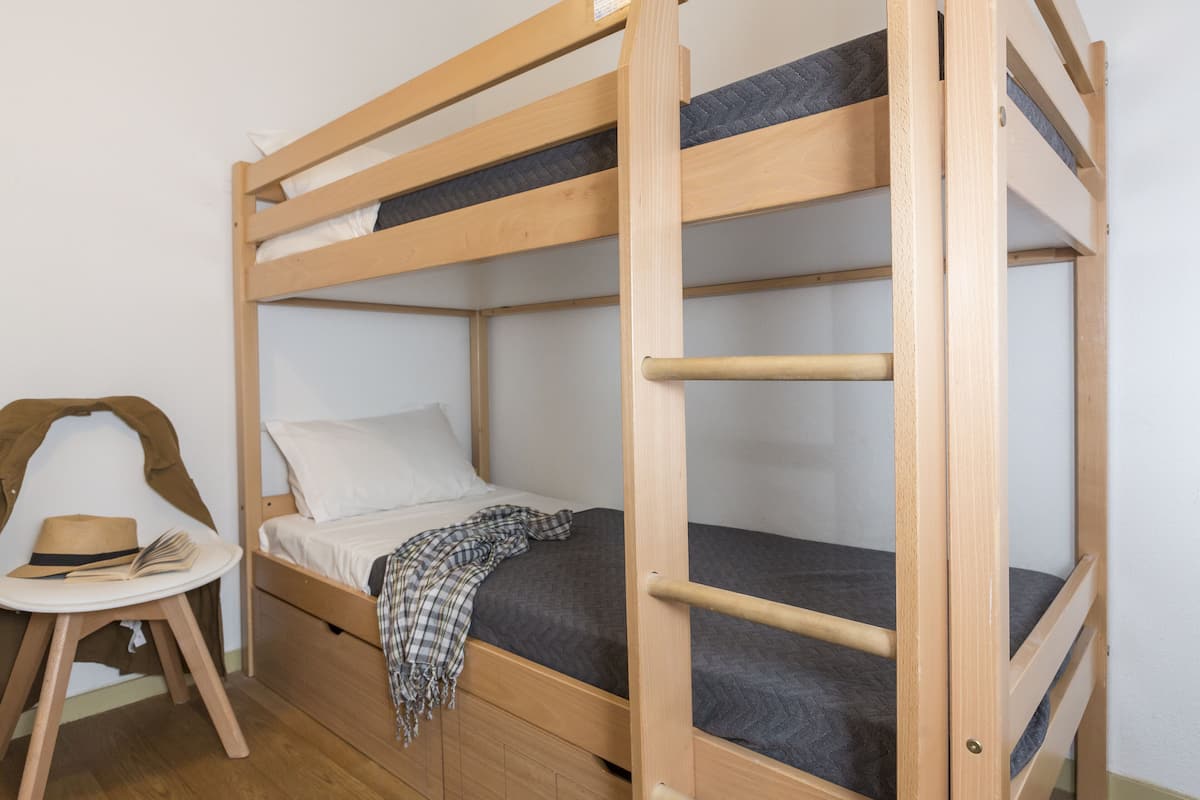 A bunk bed with a wooden frame, a small table with a hat and book, and a plaid blanket on the lower bunk.