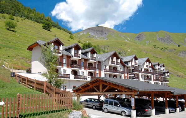 A hotel with multiple balconies, a covered parking area, and a mountainous backdrop. - Résidence Odalys L' Ouillon (Saint-Sorlin-d'Arves)