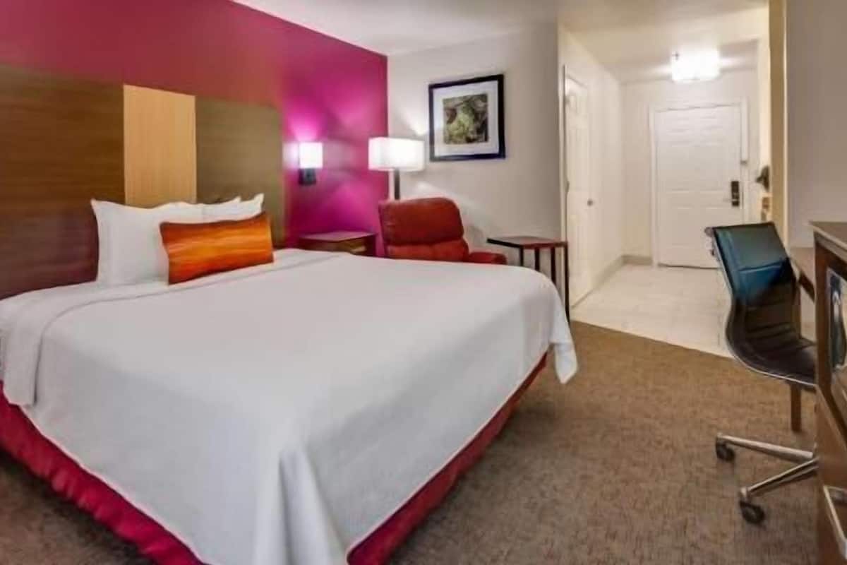 A hotel room with a large bed, a desk, a chair, a lamp, and a door.