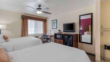A hotel room with two beds, a desk with a computer, a television, a ceiling fan, and a large mirror.