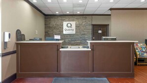 Uma área de recepção de hotel com um painel de parede de pedra, um sinal 'Quality by Choice Hotels', um balcão de recepção, uma tela digital e um letreiro com a palavra 'Bem-vindo!'.
