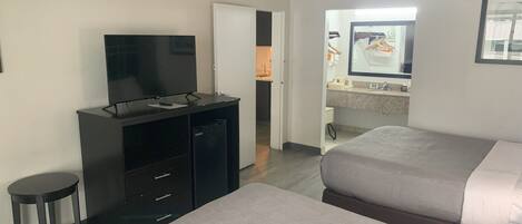 Une chambre d’hôtel avec deux lits, un téléviseur posé sur un meuble, un petit tabouret et une salle de bain visible à travers une porte ouverte.