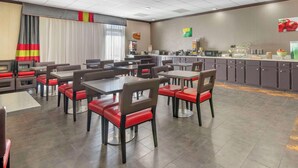 Uma área de refeições de hotel com várias mesas e cadeiras, um balcão de cozinha com equipamentos de serviço de alimentos e uma janela grande com cortinas.