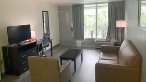 Uma sala de estar moderna com uma televisão de tela plana, um sofá, uma mesa de centro e uma poltrona. Há uma janela com cortinas, um abajur e uma pequena mesa com um abajur.