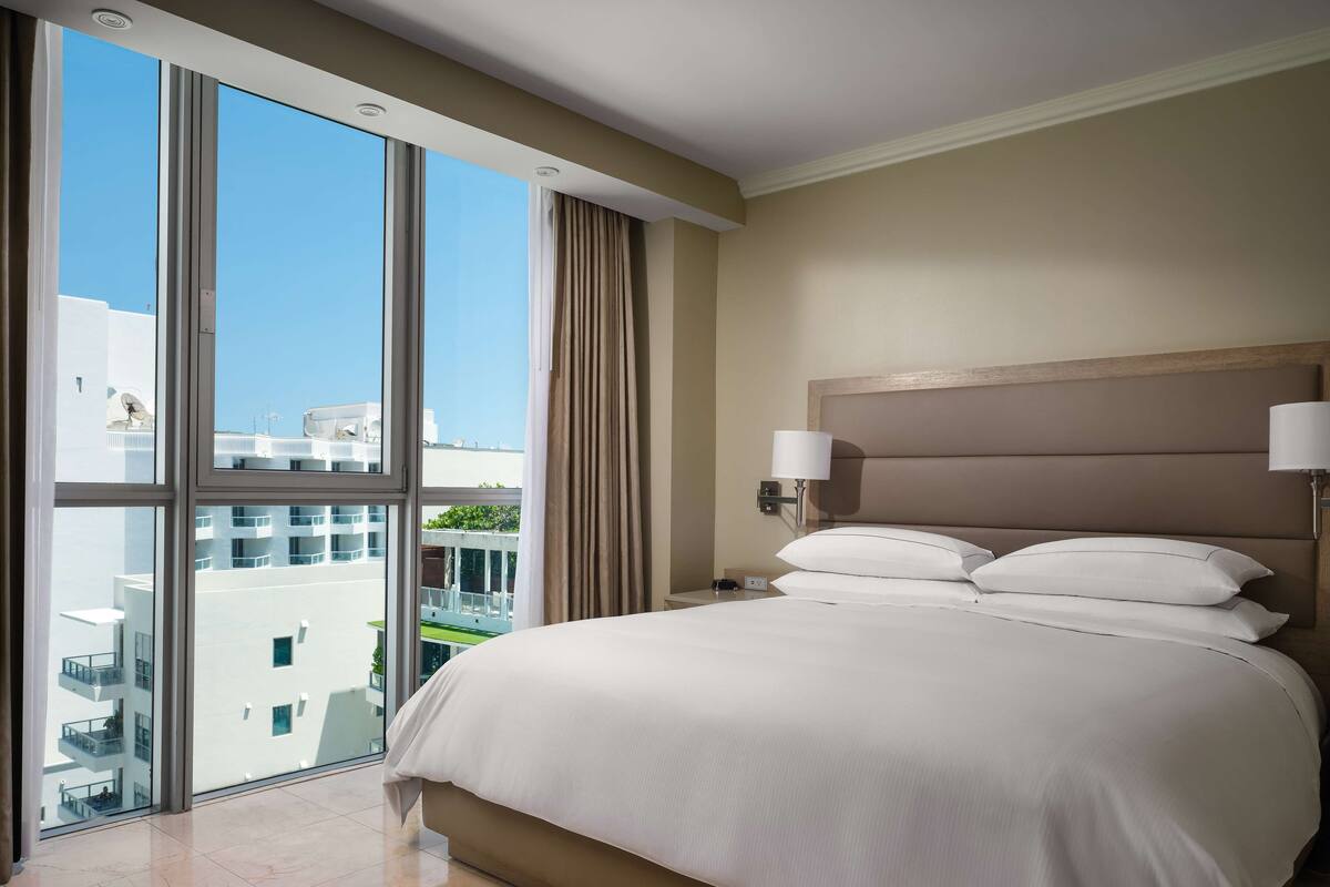 A hotel room with a large bed, two bedside lamps, a view of the city through floor-to-ceiling windows, and a modern headboard.