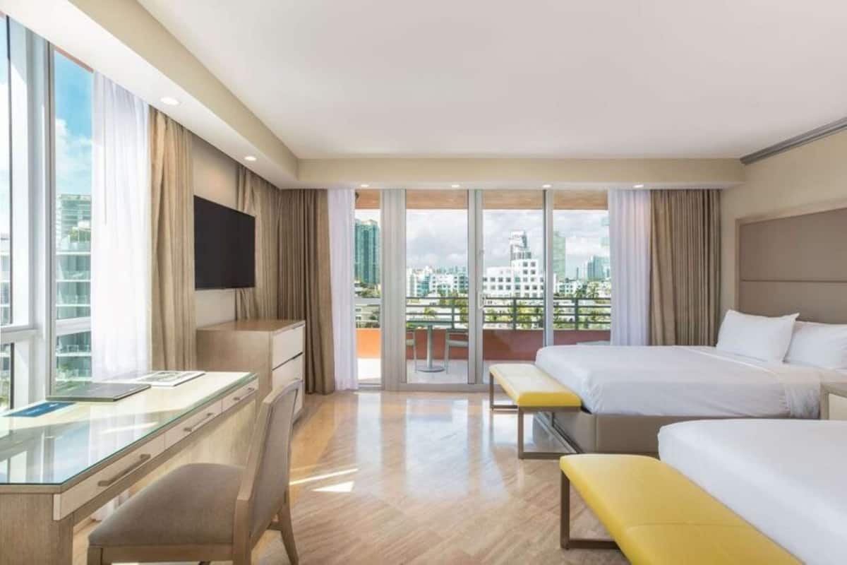 Habitación de hotel con dos camas, un escritorio, una silla, un televisor y un balcón con vistas a la ciudad.