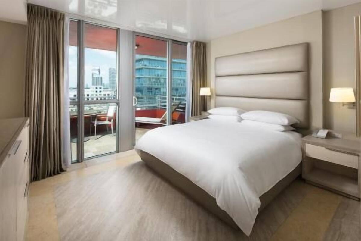 Una habitación de hotel con una cama grande, una mesita de noche, una ventana con cortinas y vistas a un edificio.