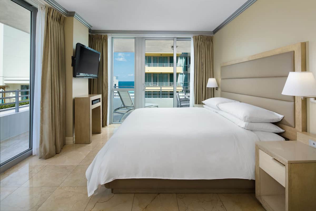 Un cuarto de hotel con una cama grande, un televisor montado en la pared, un balcón con vista al mar y un escritorio con una lámpara.
