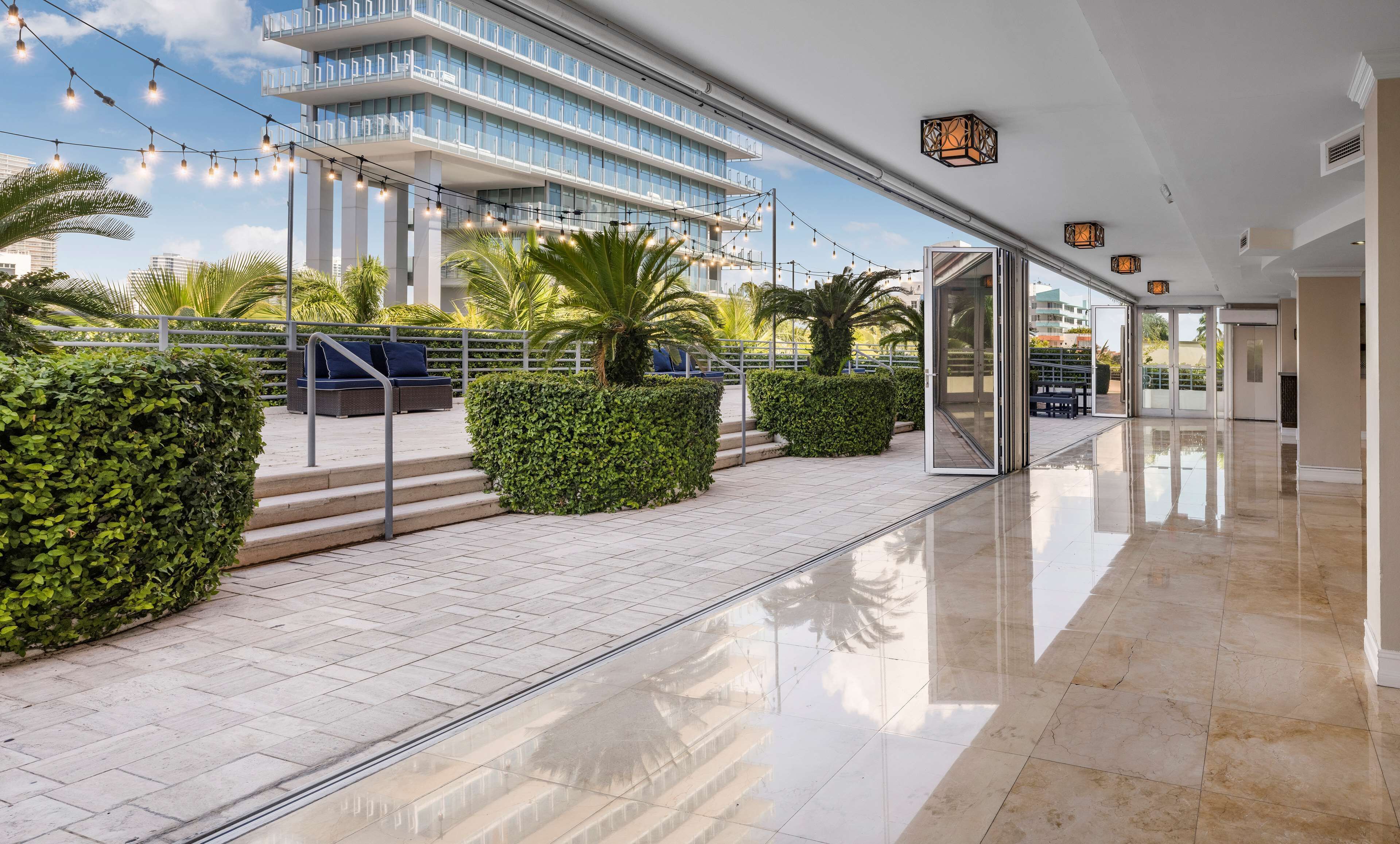 A modern hotel lobby with a glass entrance, a tiled floor, and outdoor seating with palm trees.