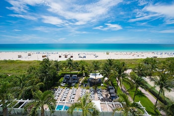 A hotel room with a large bed, a TV mounted on the wall, a balcony with a view of the beach, and a small table with a chair. at Hilton Bentley Miami/South Beach