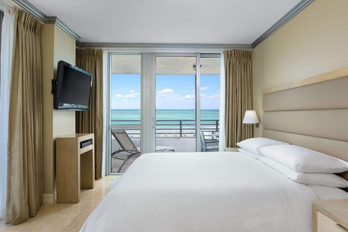 A hotel room with a large bed, a TV mounted on the wall, a balcony with a view of the sea, and a desk with a lamp.