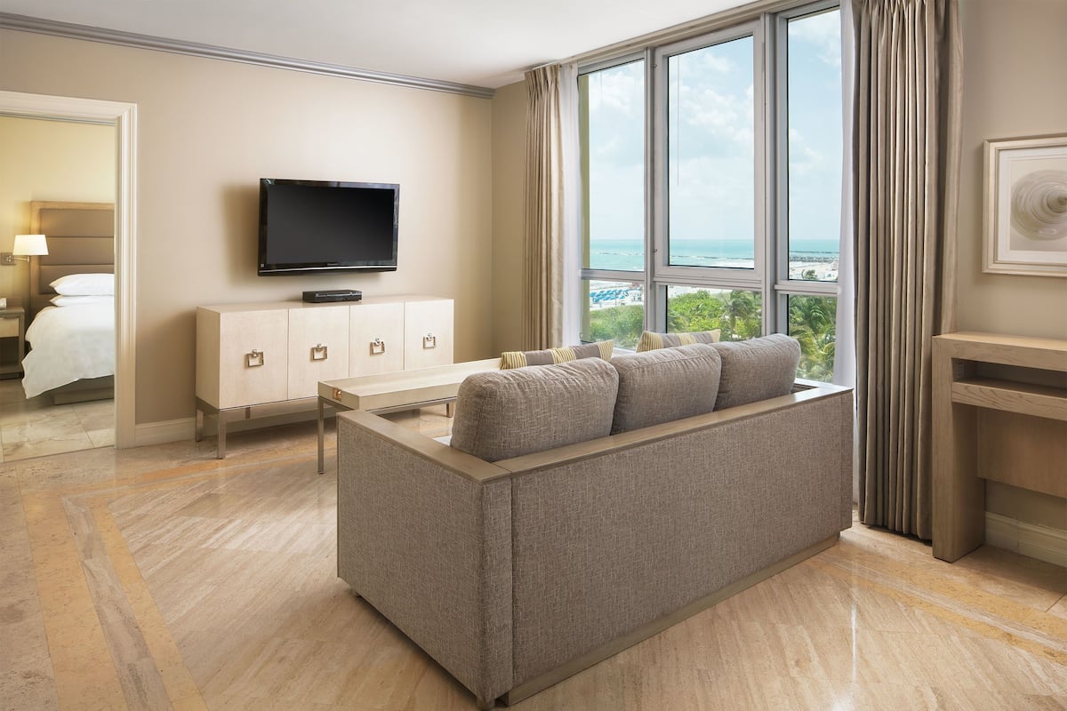 Habitación de hotel moderna con sofá, televisor, mueble y ventana grande con vista al océano.