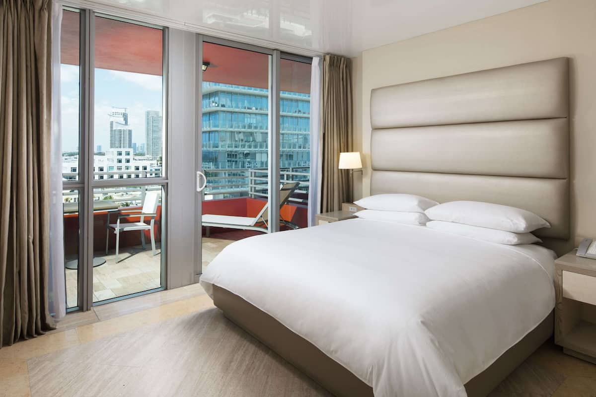 Habitación de hotel con una cama grande, una mesita de noche, una lámpara y vistas a la ciudad a través de una puerta corrediza de vidrio.
