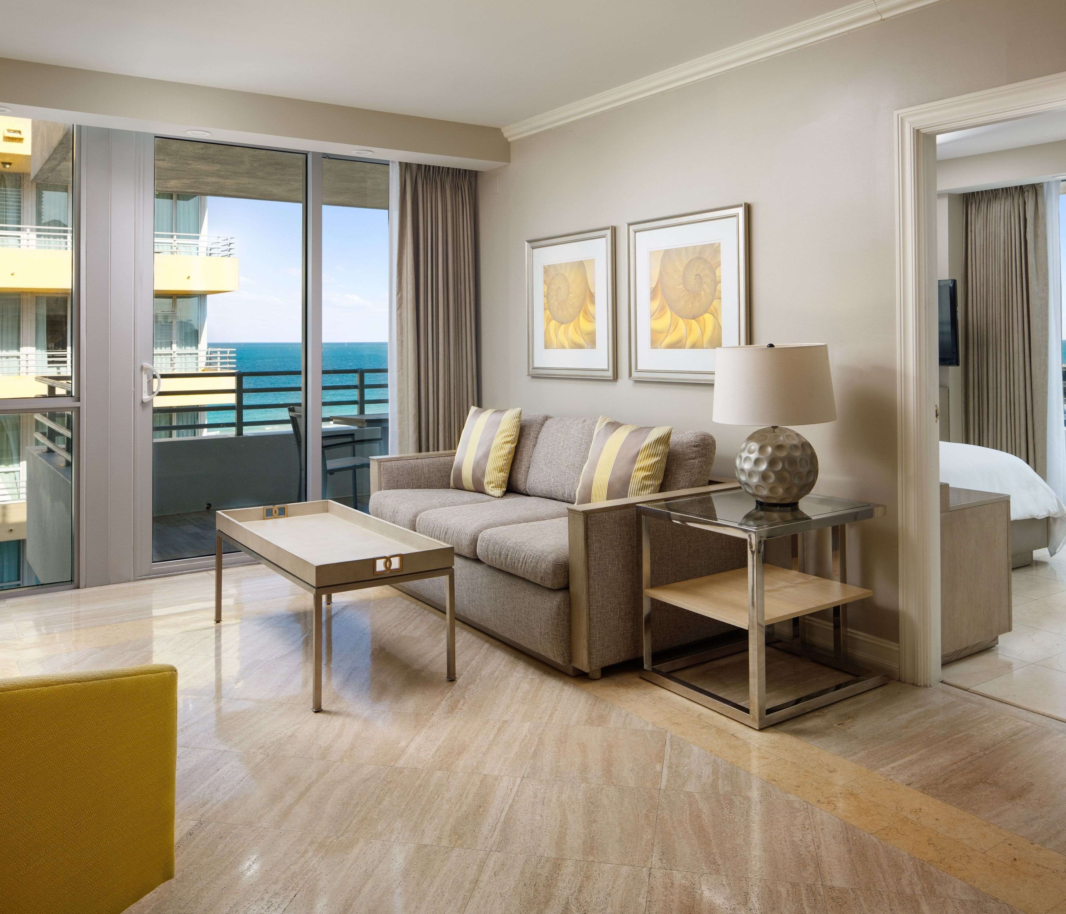 A modern hotel room with a sofa, a side table with a lamp, a glass coffee table, and a bed.