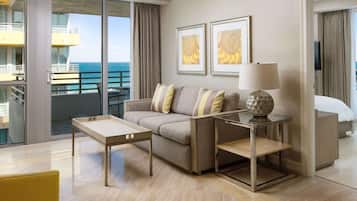 A modern hotel room with a sofa, a side table with a lamp, a glass coffee table, and a bed.