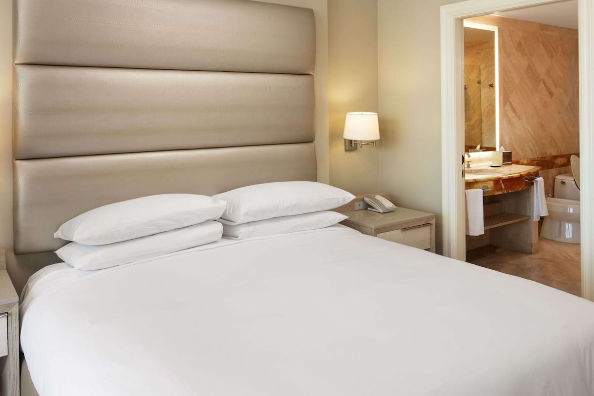 Habitación de hotel con una cama grande, una mesita de noche con lámpara y un baño visible a través de una puerta abierta.
