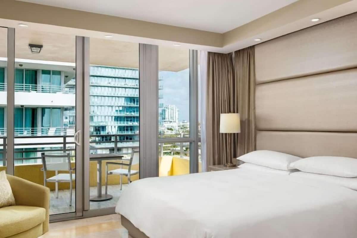 Habitación de hotel con una cama grande, un sofá, un escritorio y vistas a un paisaje urbano a través de grandes ventanales.