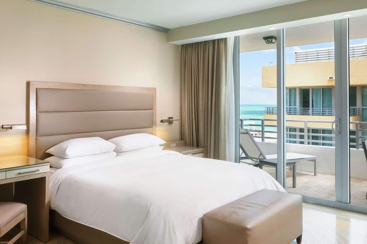 Habitación de hotel con una cama grande, una mesita de noche, una silla y vistas al océano a través de amplias ventanas.