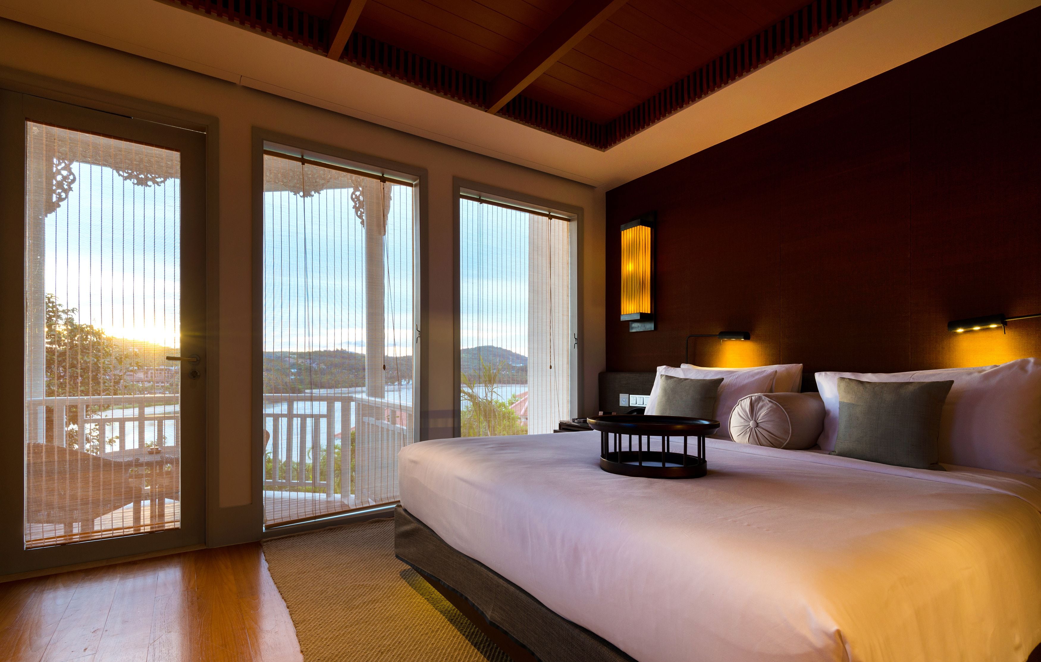 Una cama grande con cabecera de madera, una mesita de noche con lámpara, y vistas al exterior a través de puertas corredizas de vidrio.