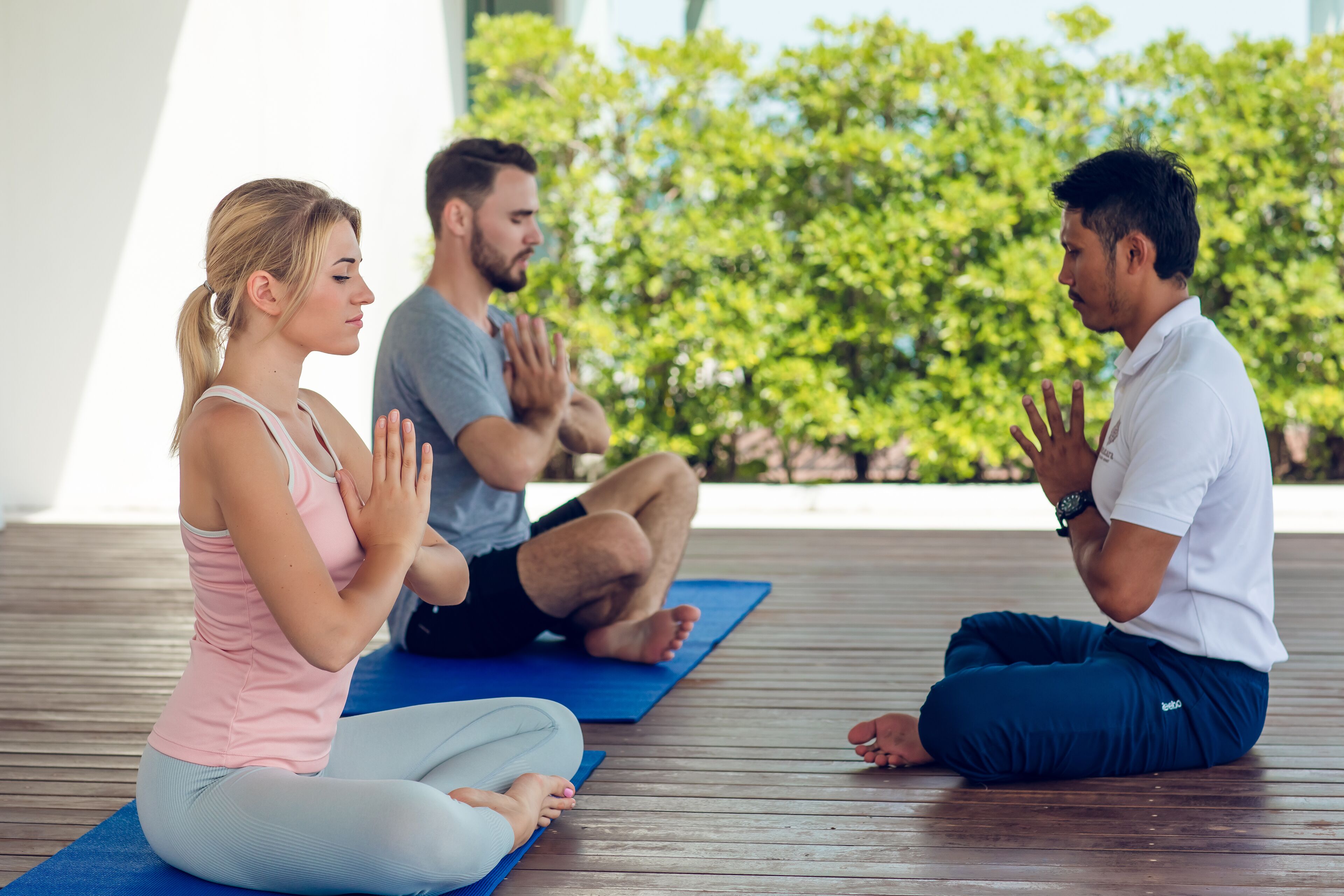 Three people sitting on yoga mats in an outdoor setting with a wooden deck and greenery in the background.