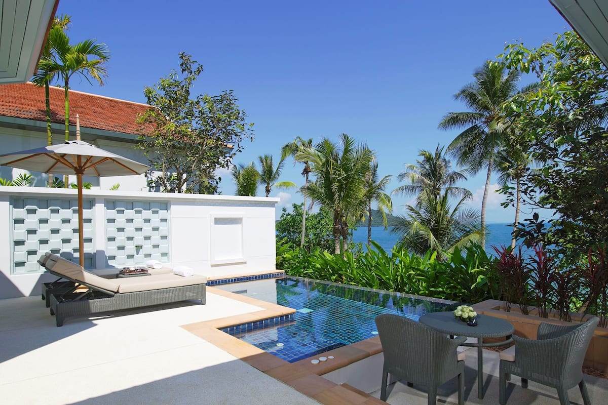 A pool area with lounge chairs and a table, surrounded by tropical plants and a view of the ocean.