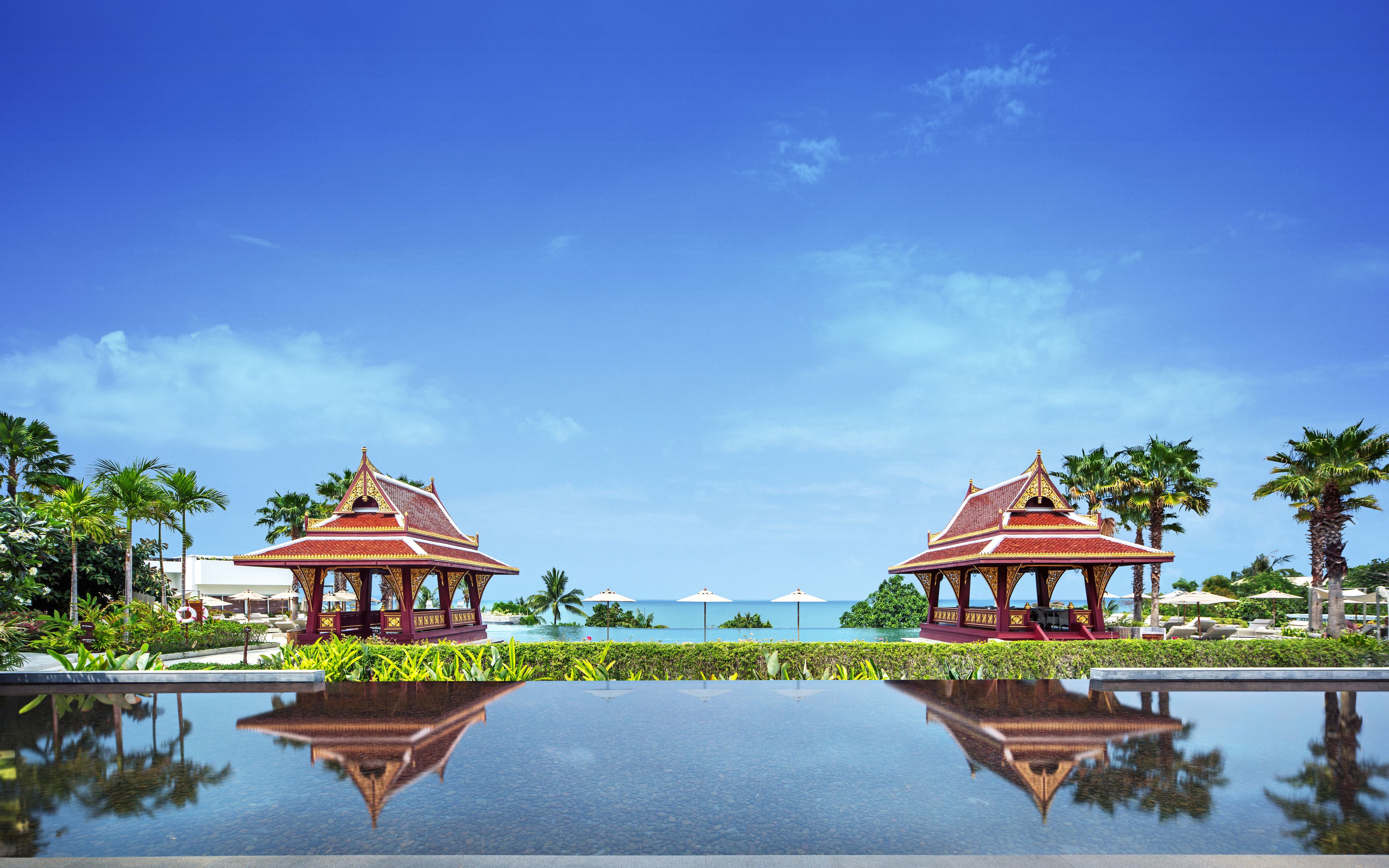 Ein Resort mit zwei Gazebos mit roten Dächern, blauem Himmel und einem ruhigen Pool, der die Szene spiegelt.