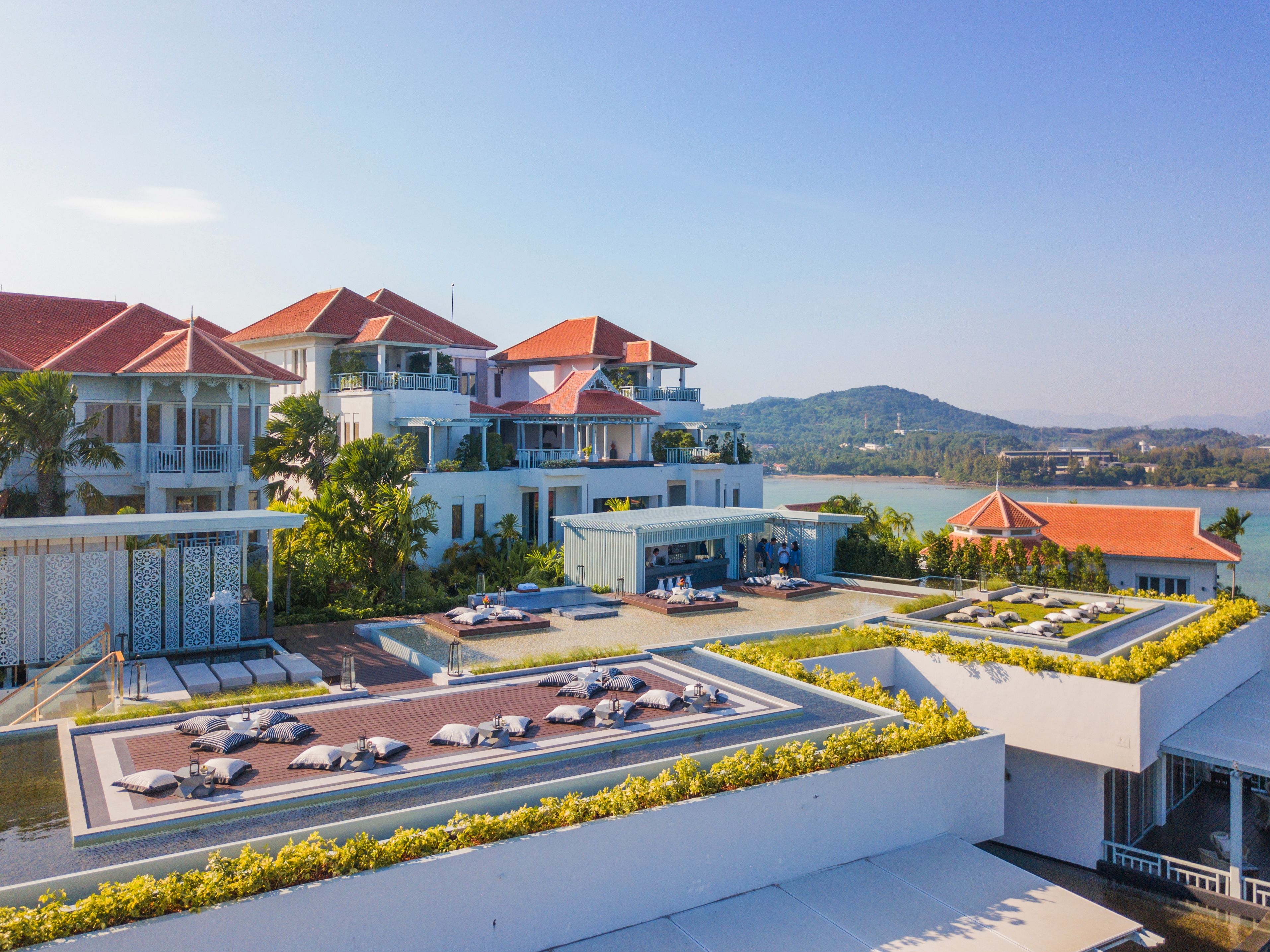 A resort with multiple buildings, a rooftop lounge area with sun loungers and umbrellas, and a view of the sea and hills in the background.