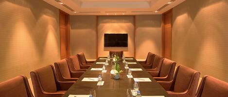 A conference room with a long table, brown upholstered chairs, and a flat-screen TV on the wall.