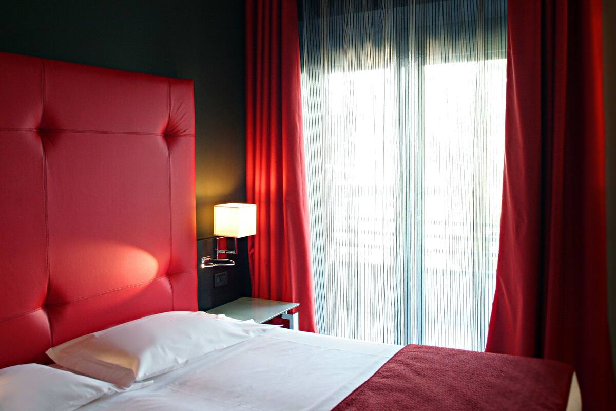 A hotel room with a large red headboard, a bed with white linens, a nightstand with a lamp, and a window with red curtains.