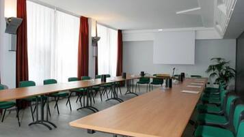Un salón de conferencias con una larga mesa, sillas verdes, una pantalla de proyector y una planta.