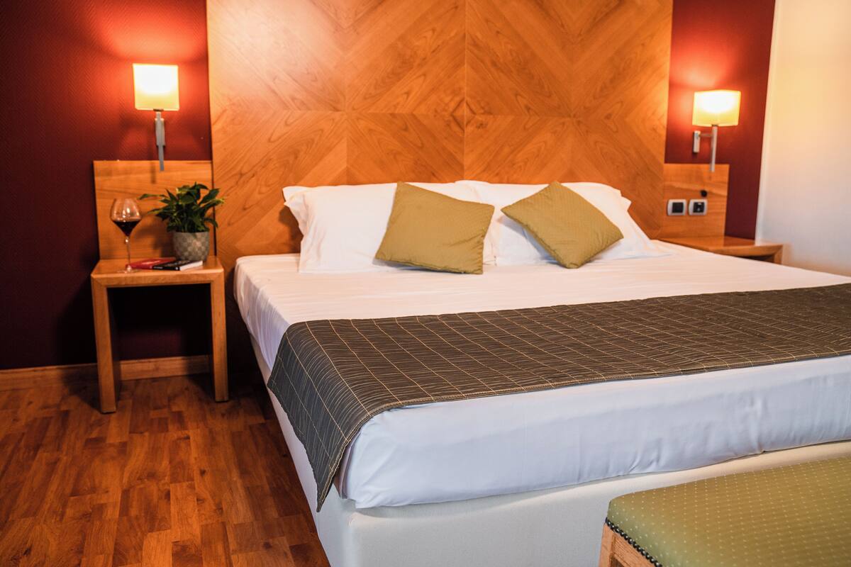 A hotel room with a wooden headboard, a bed with white linens and two pillows, a bedside table with a lamp, a small plant, and a wine glass.