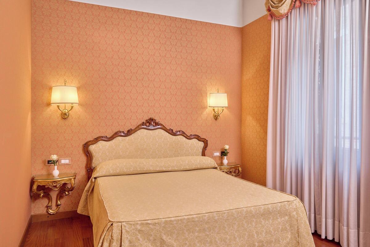 Habitación de hotel con cama, dos lámparas de noche, mesita de noche con un florero y ventana con cortinas.