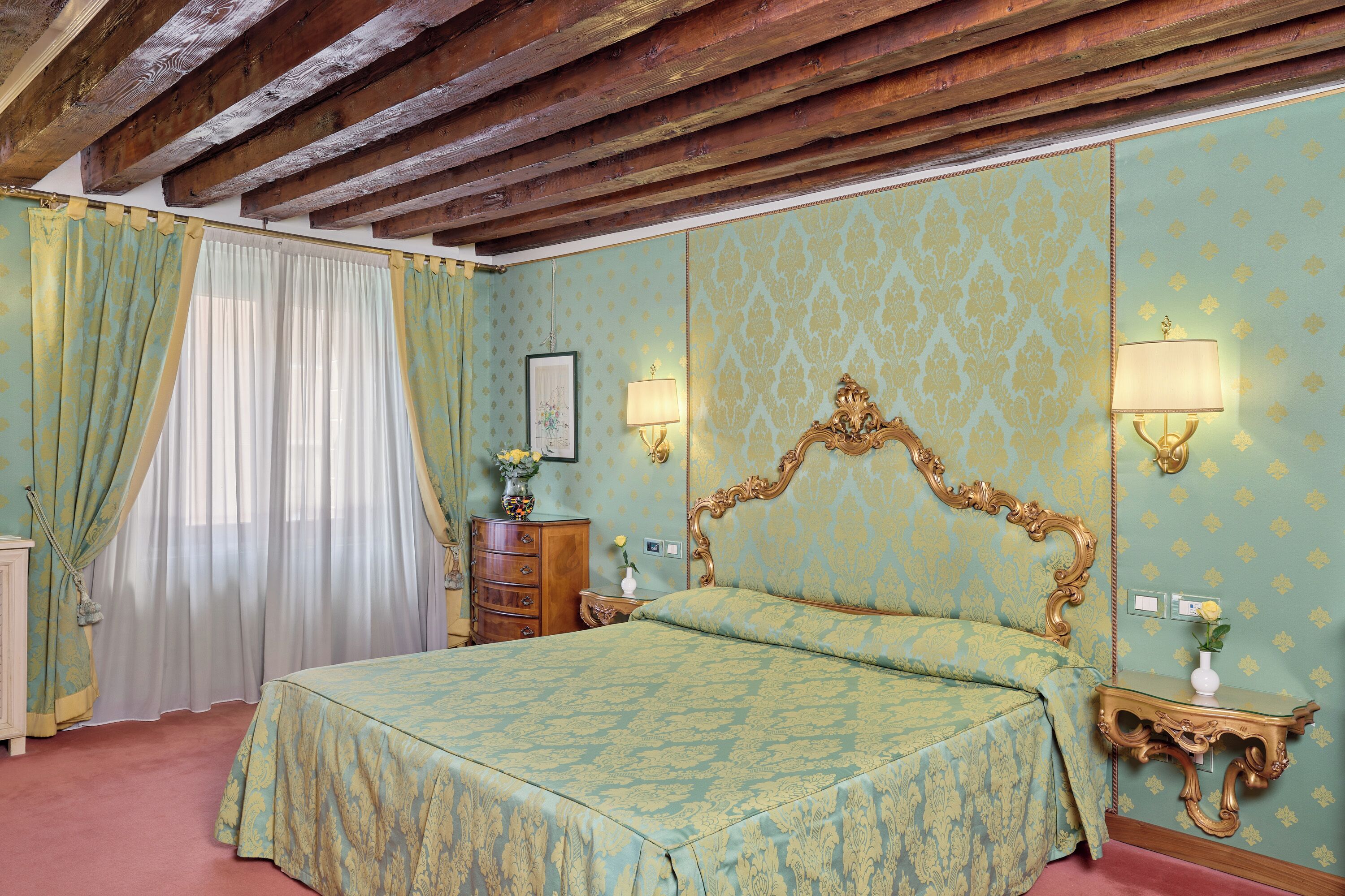 Quarto com cama, vigas de madeira no teto, penteadeira, duas luminárias na parede e um vaso com flores.