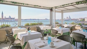 Una terraza con mesas dispuestas para comer, con vistas a un paisaje urbano que incluye edificios destacados y una masa de agua.