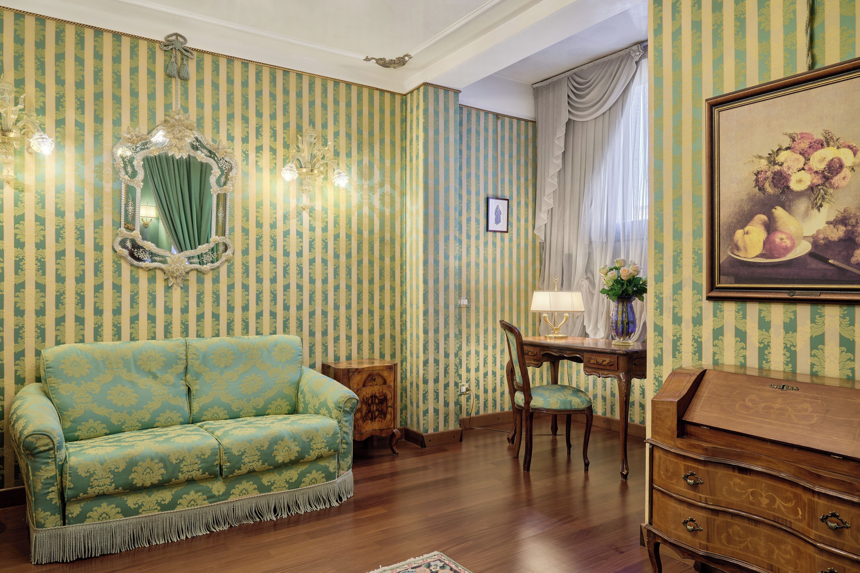 A room with a patterned wallpaper, a green sofa, a wooden dresser, a dining table with a chair, and a chandelier.