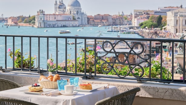 Un balcón con una mesa lista para el desayuno, con vista a un muelle con barcos y edificios históricos.
