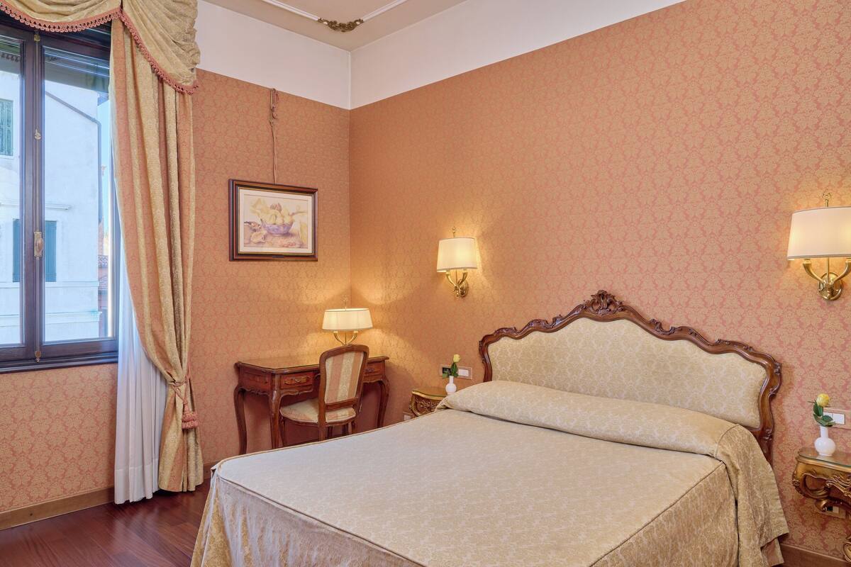 Una habitación con cama, escritorio, silla, dos lámparas de pared y un cuadro enmarcado.