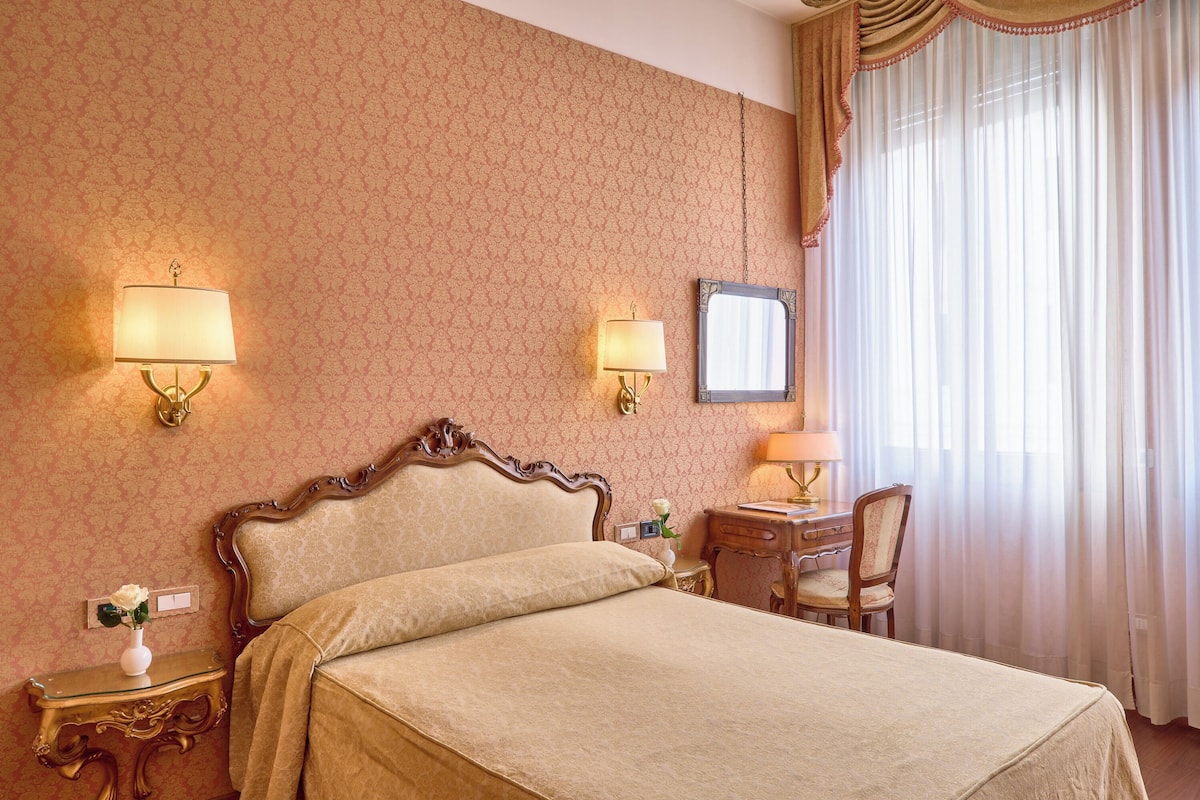 Habitación de hotel con cama, dos lámparas de noche, escritorio con silla y ventana grande con cortinas.