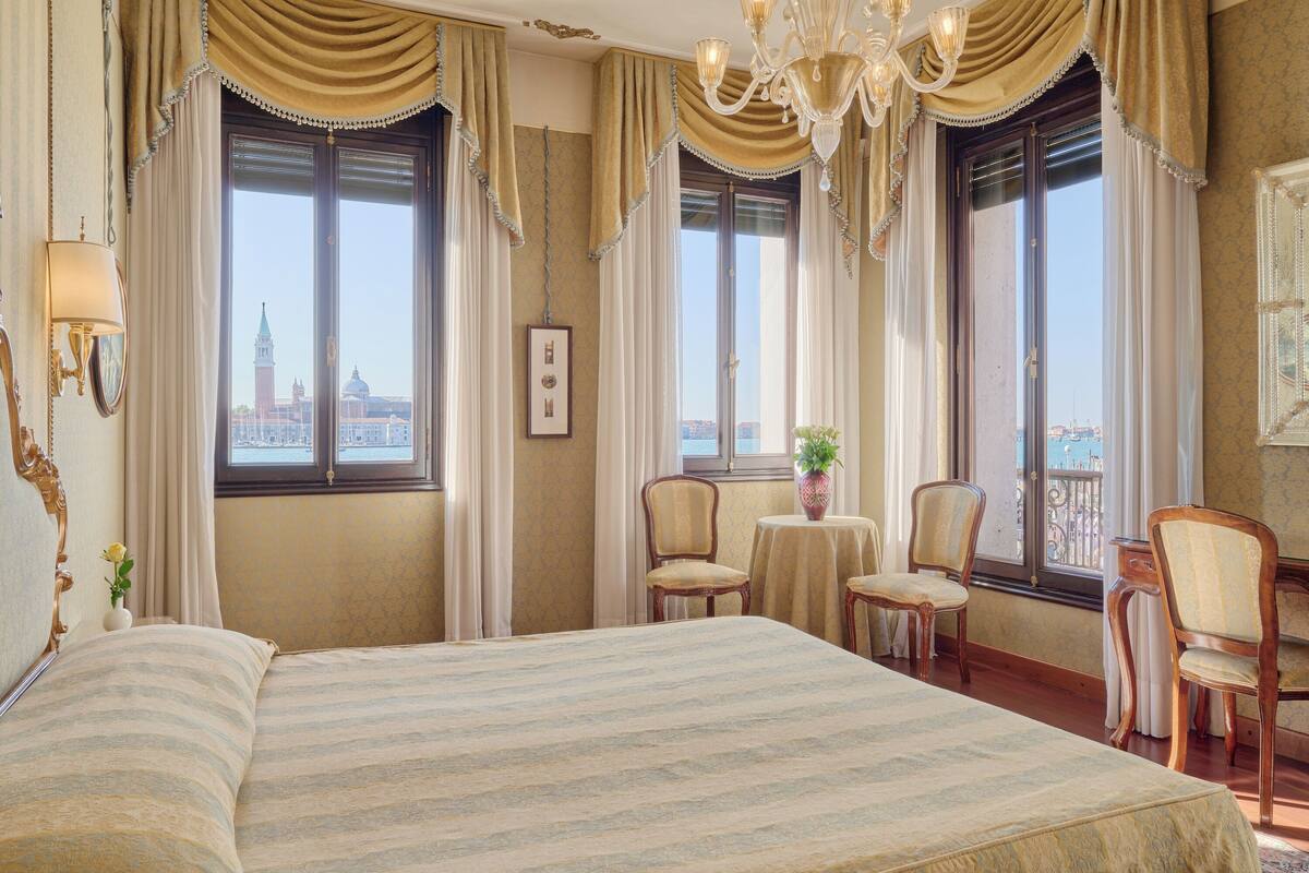 Habitación de hotel con una cama grande, dos sillas, una mesita con una planta y vistas a la ciudad a través de las ventanas.