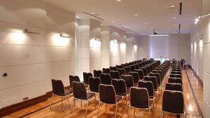 Un salón de conferencias con filas de sillas, piso de madera y paredes blancas con iluminación empotrada.